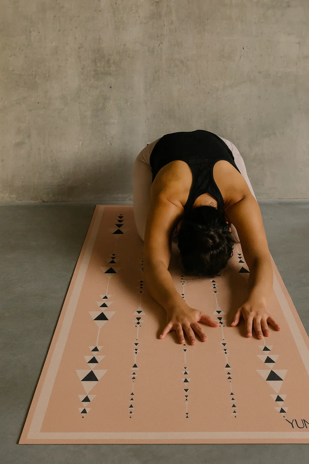 Person practicing yoga on a pink mat with black triangle patterns against a concrete wall.