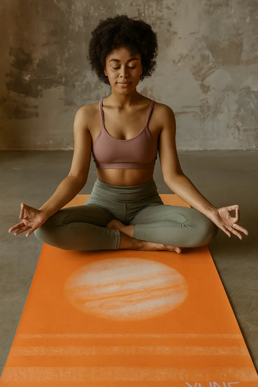 Woman meditating on an orange yoga mat with a Jupiter design against a textured wall.