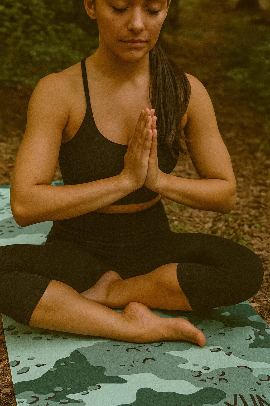 Woman in black athletic wear sitting in a yoga pose on a camouflage mat outdoors.