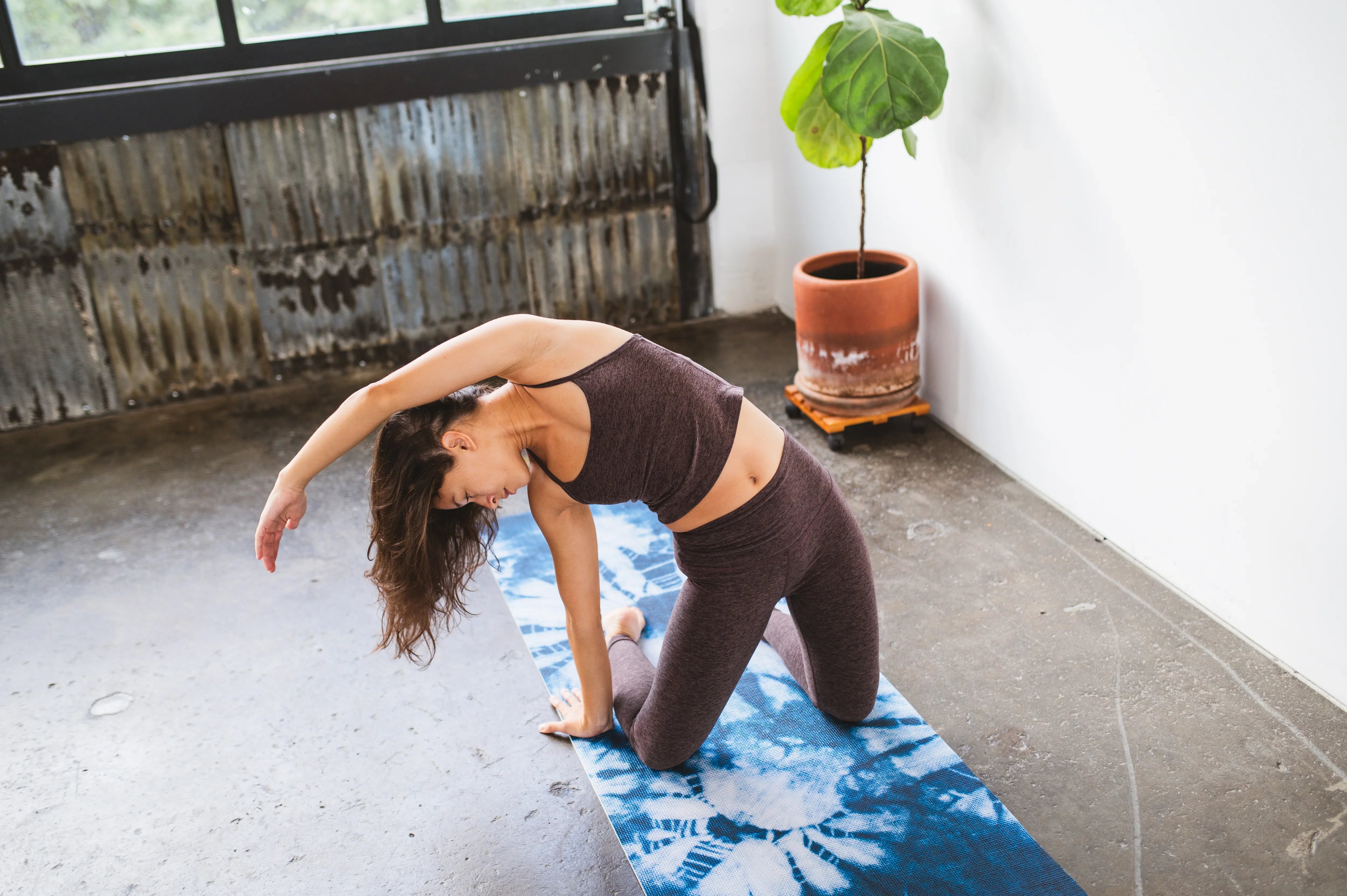 Woman practicing yoga on a blue mat in a room with a plant and metal wall.