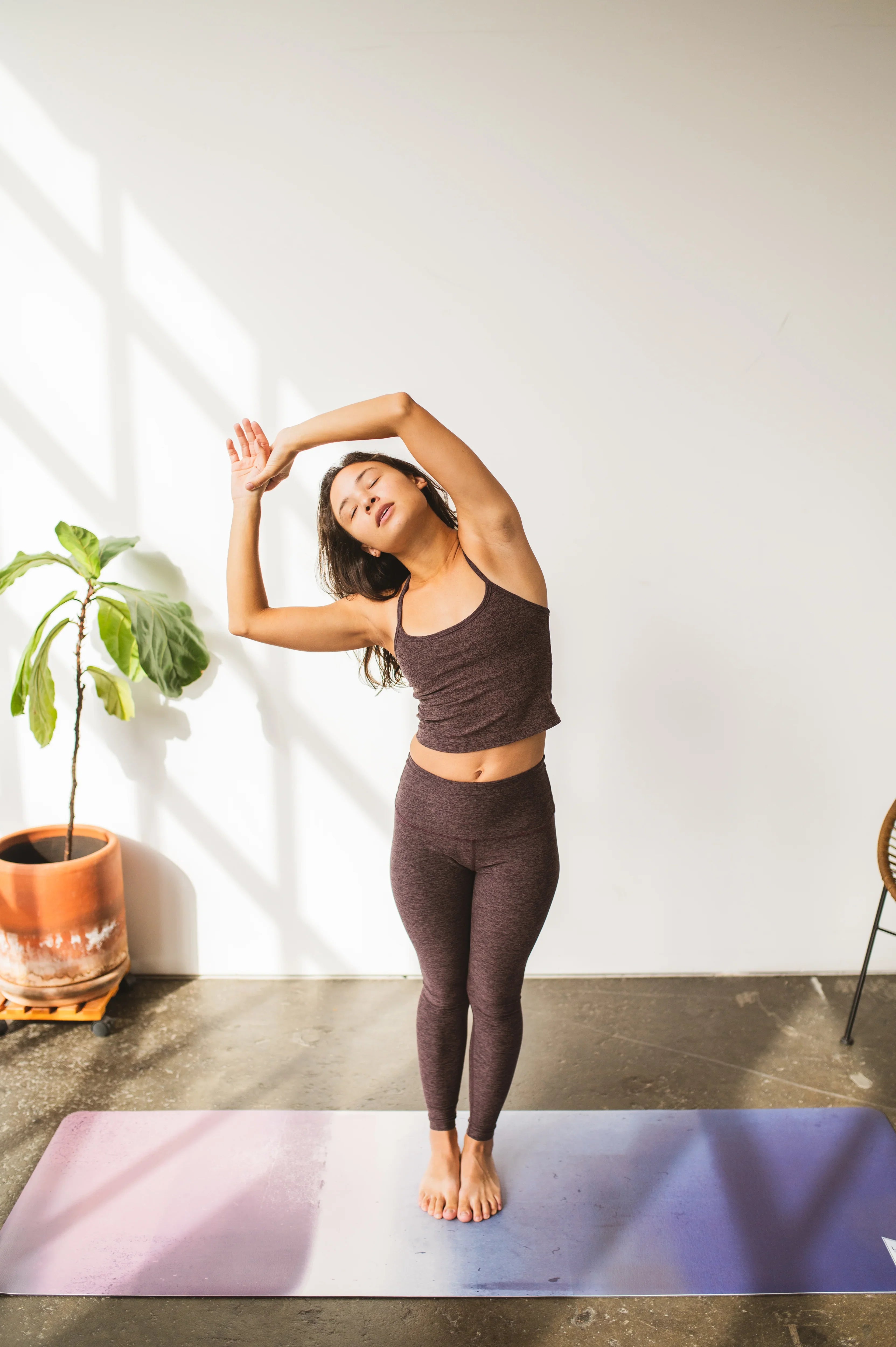 Woman in brown athletic wear stretching on a yoga mat with a plant and chair in the background.