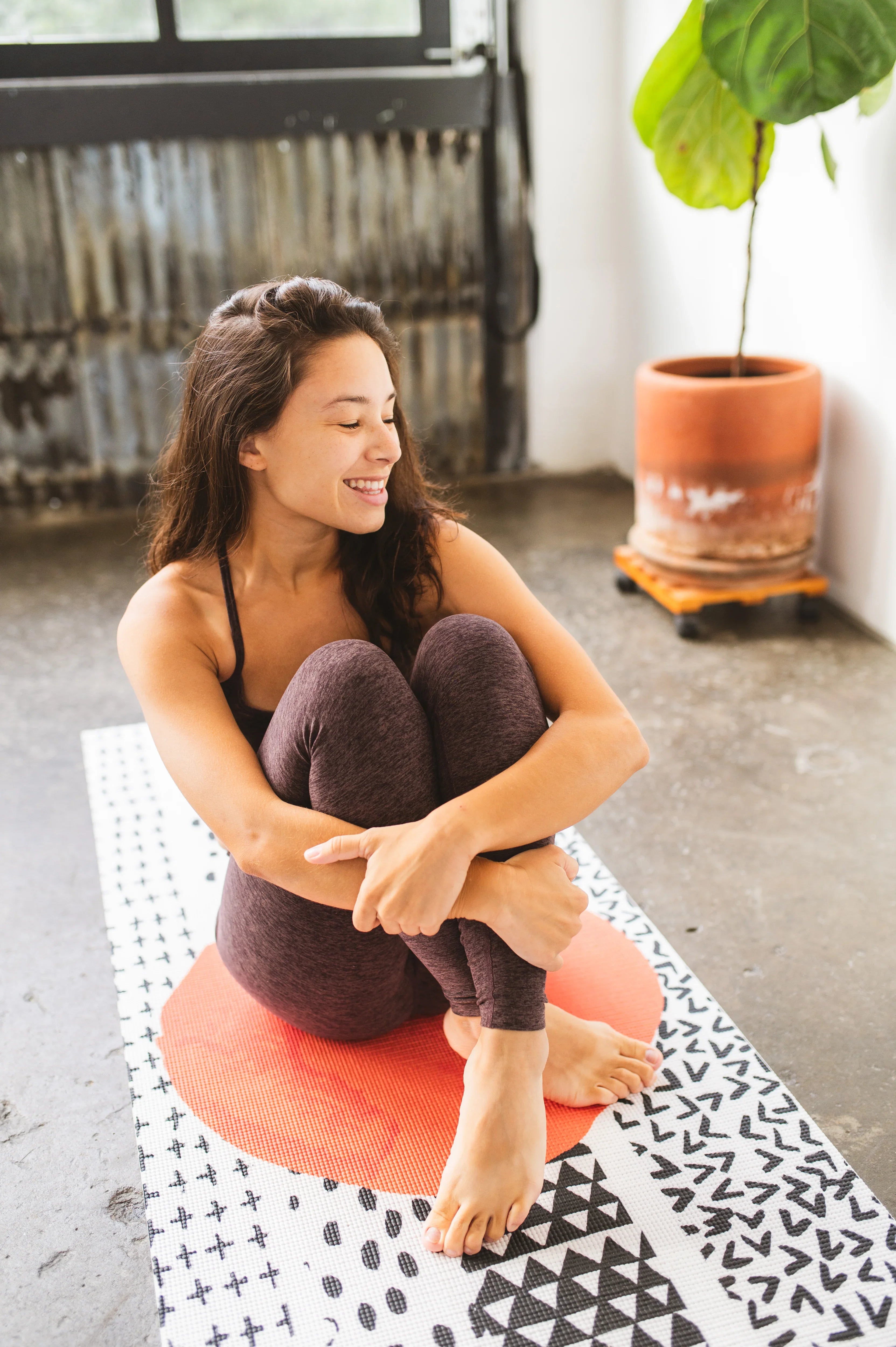 Woman sitting on a pink yoga bolster in a room with a plant and patterned rug.