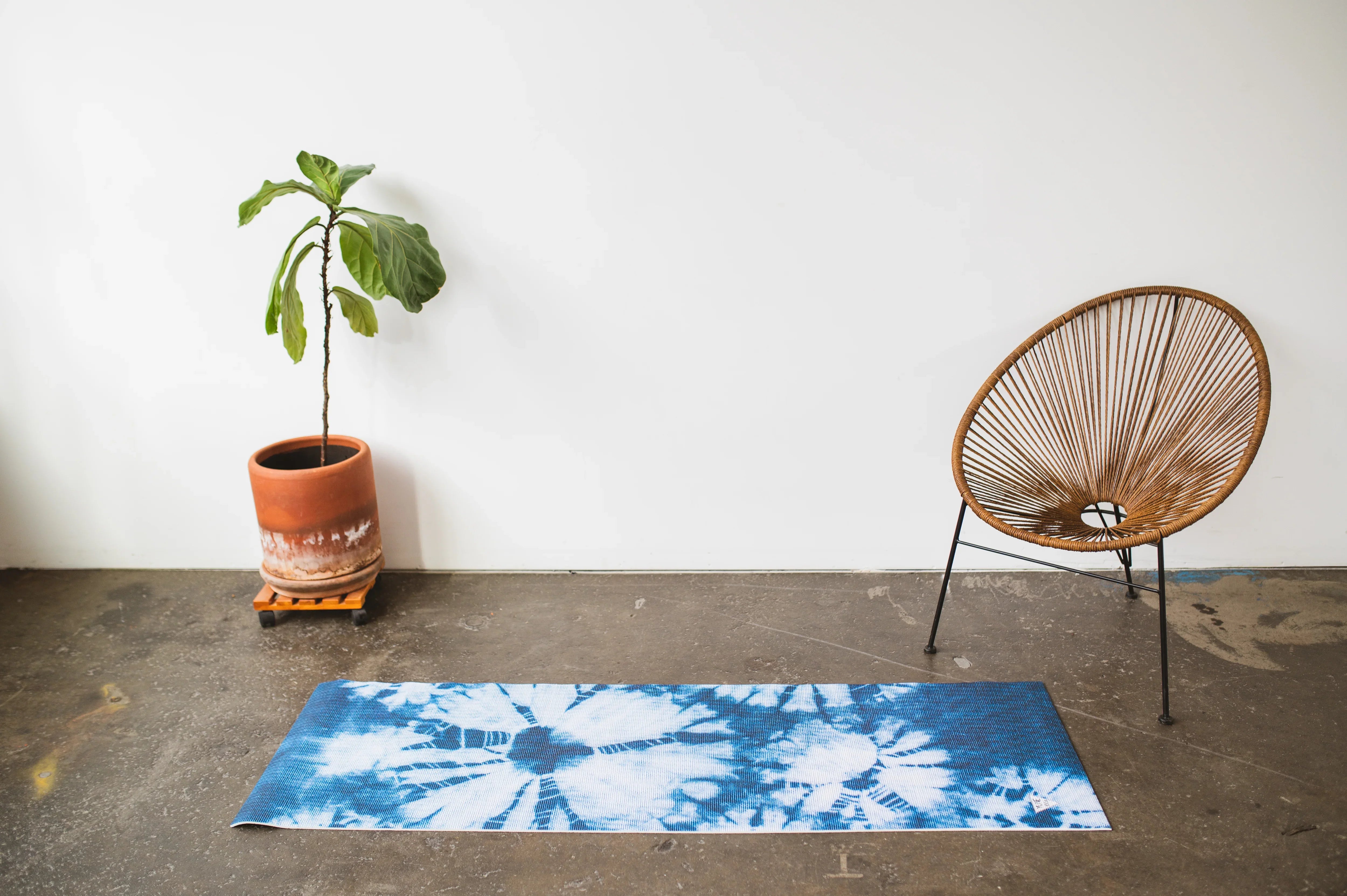 Yoga mat with tie-dye pattern on a concrete floor, next to a potted plant and a woven chair.