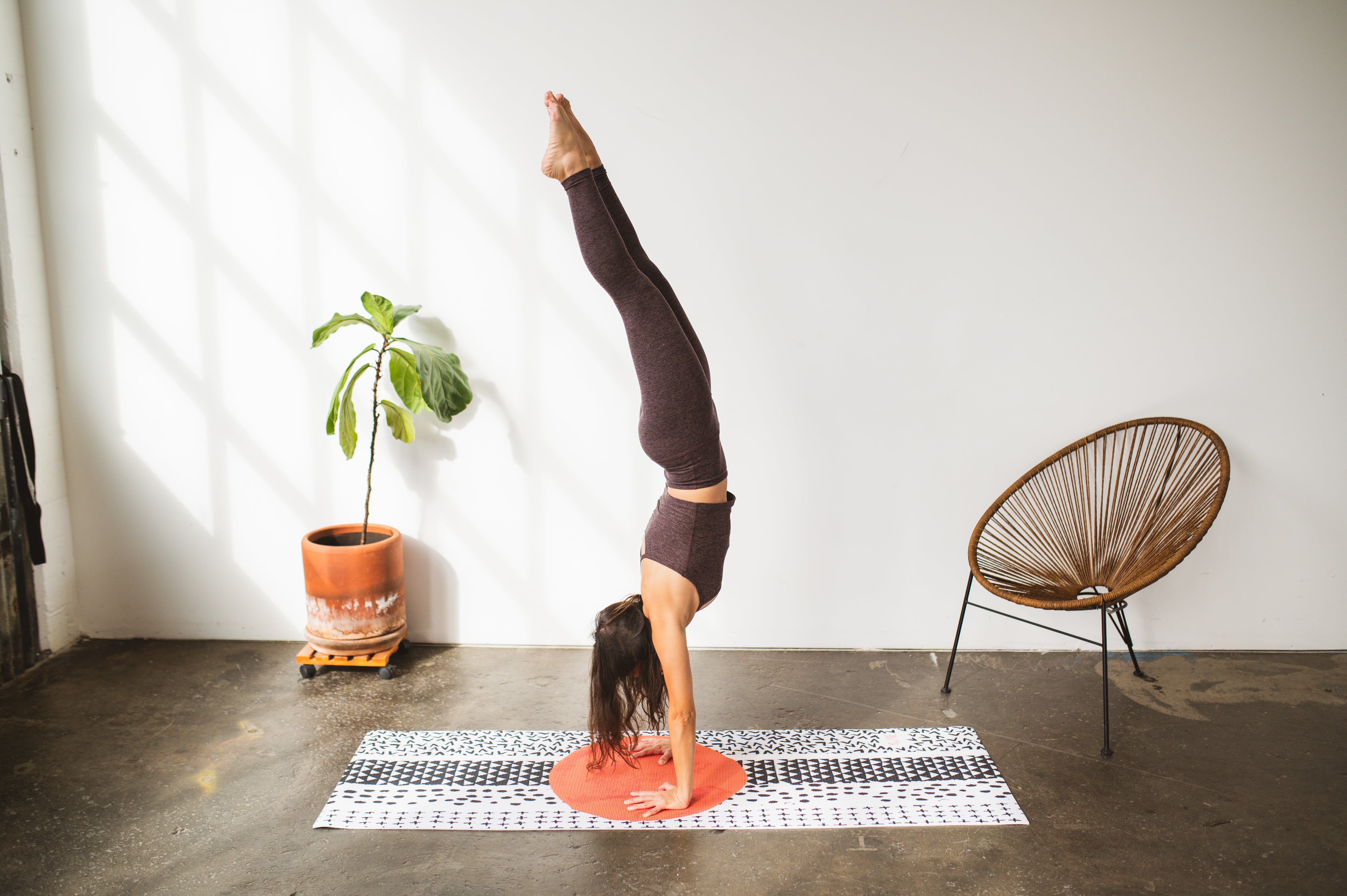 Person performing a handstand on a yoga mat in a room with a plant and chair.