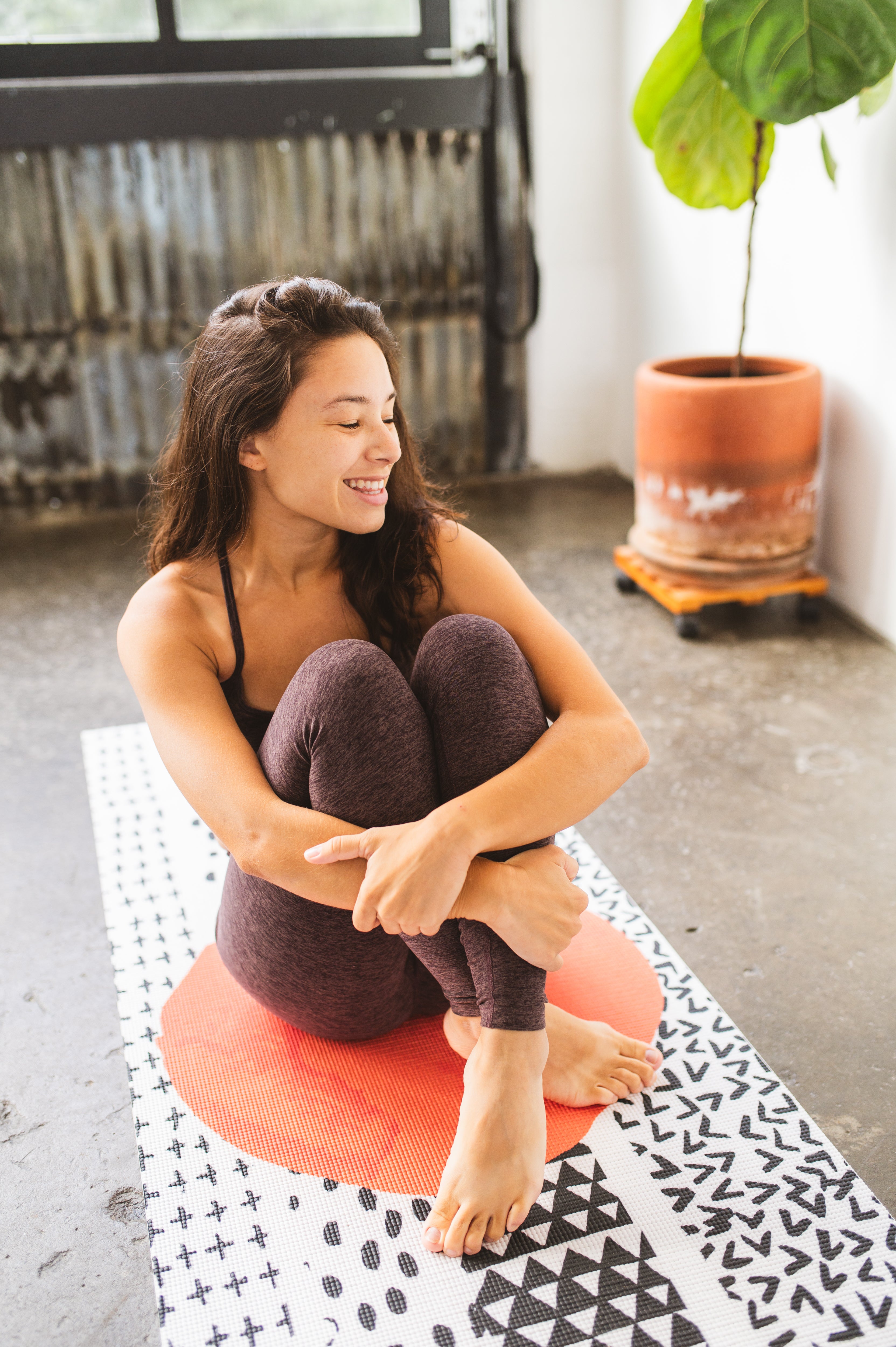Woman sitting on a pink yoga bolster in a room with a plant and patterned rug.