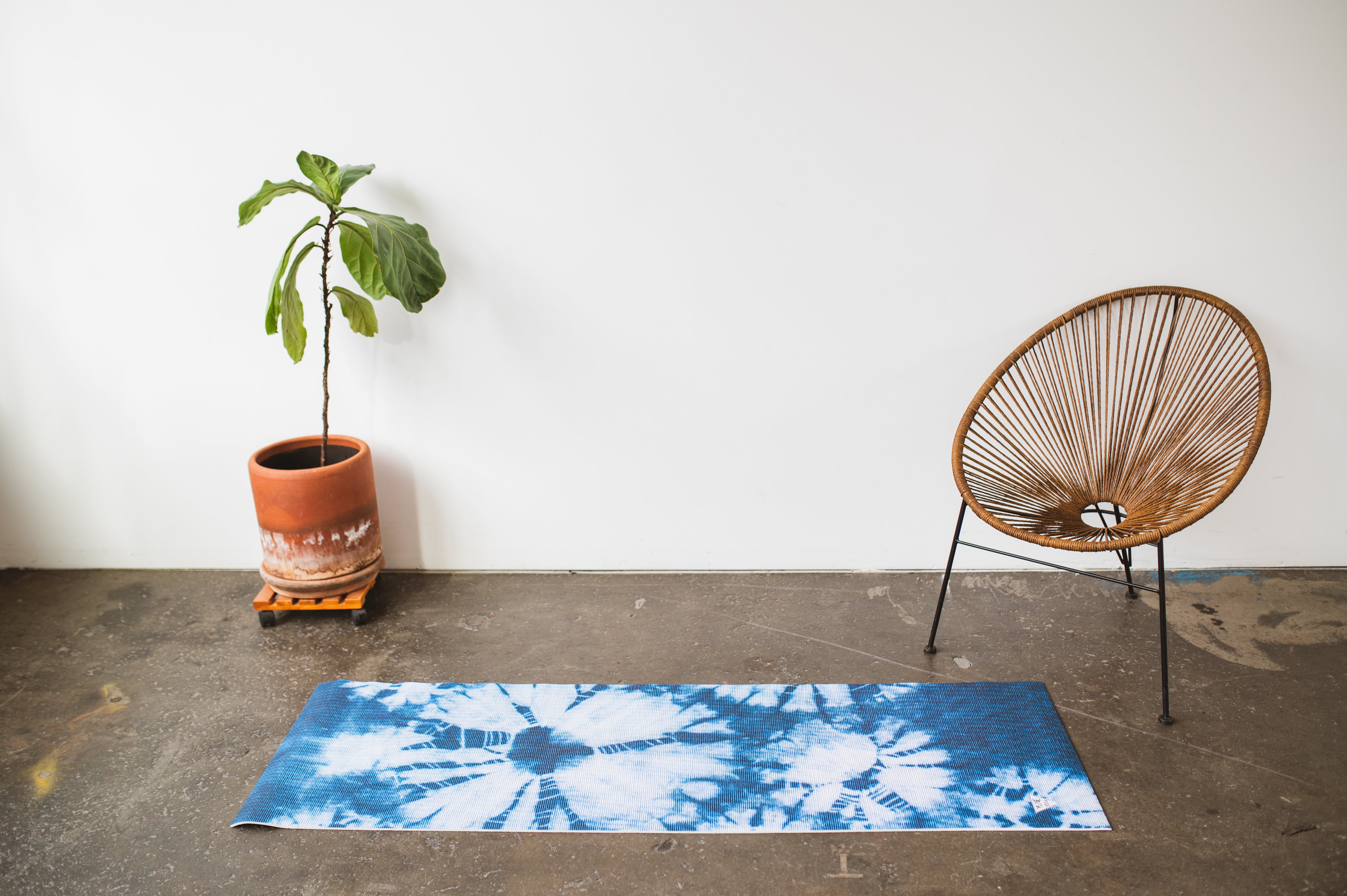 Yoga mat with tie-dye pattern on a concrete floor, next to a potted plant and a woven chair.