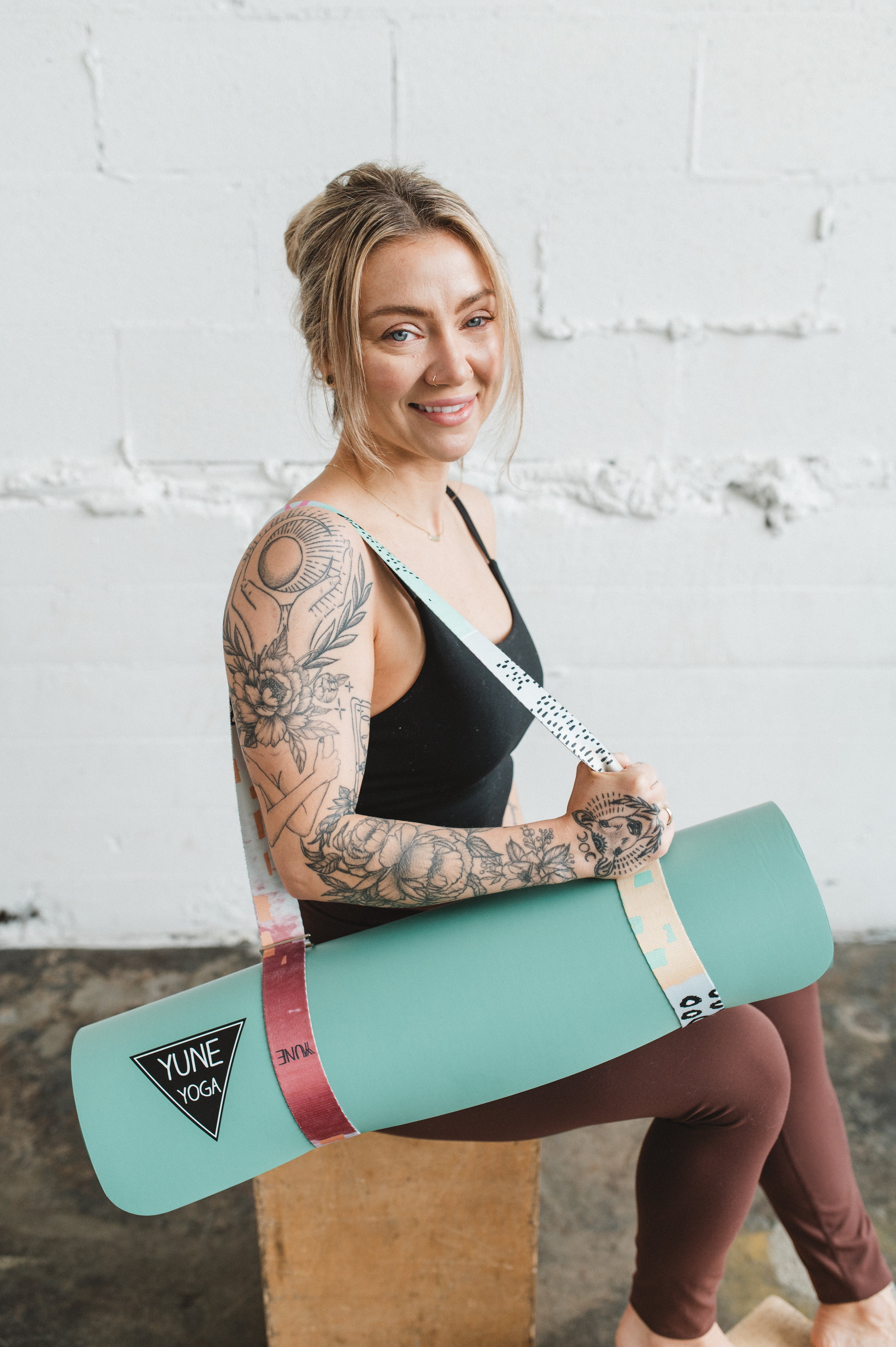 Woman holding a teal Yuna Yoga mat against a white brick wall.